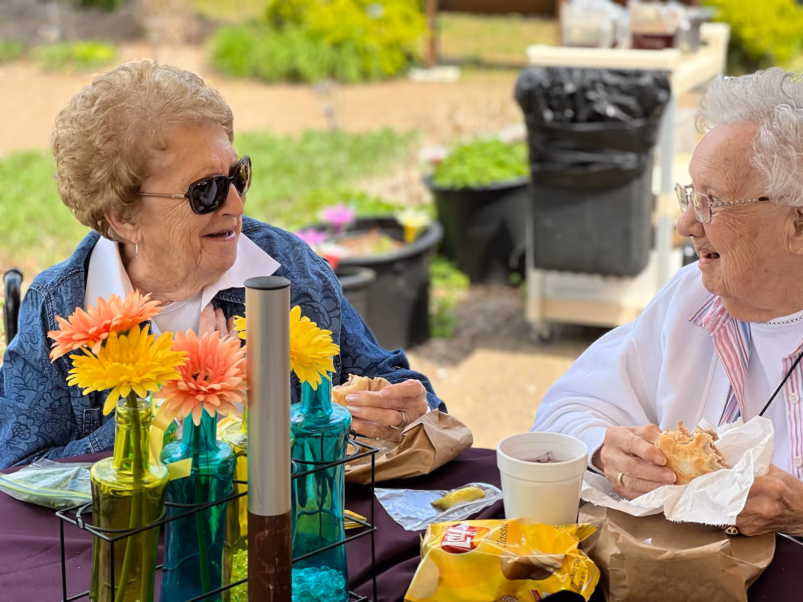 Two elderly women sitting at a table outdoors, enjoying a meal together. One woman is wearing sunglasses and a blue jacket, while the other wears glasses and a white sweater. On the table are colorful flowers in glass bottles, a bag of Lay's Classic potato chips, a white cup, and sandwiches wrapped in paper.