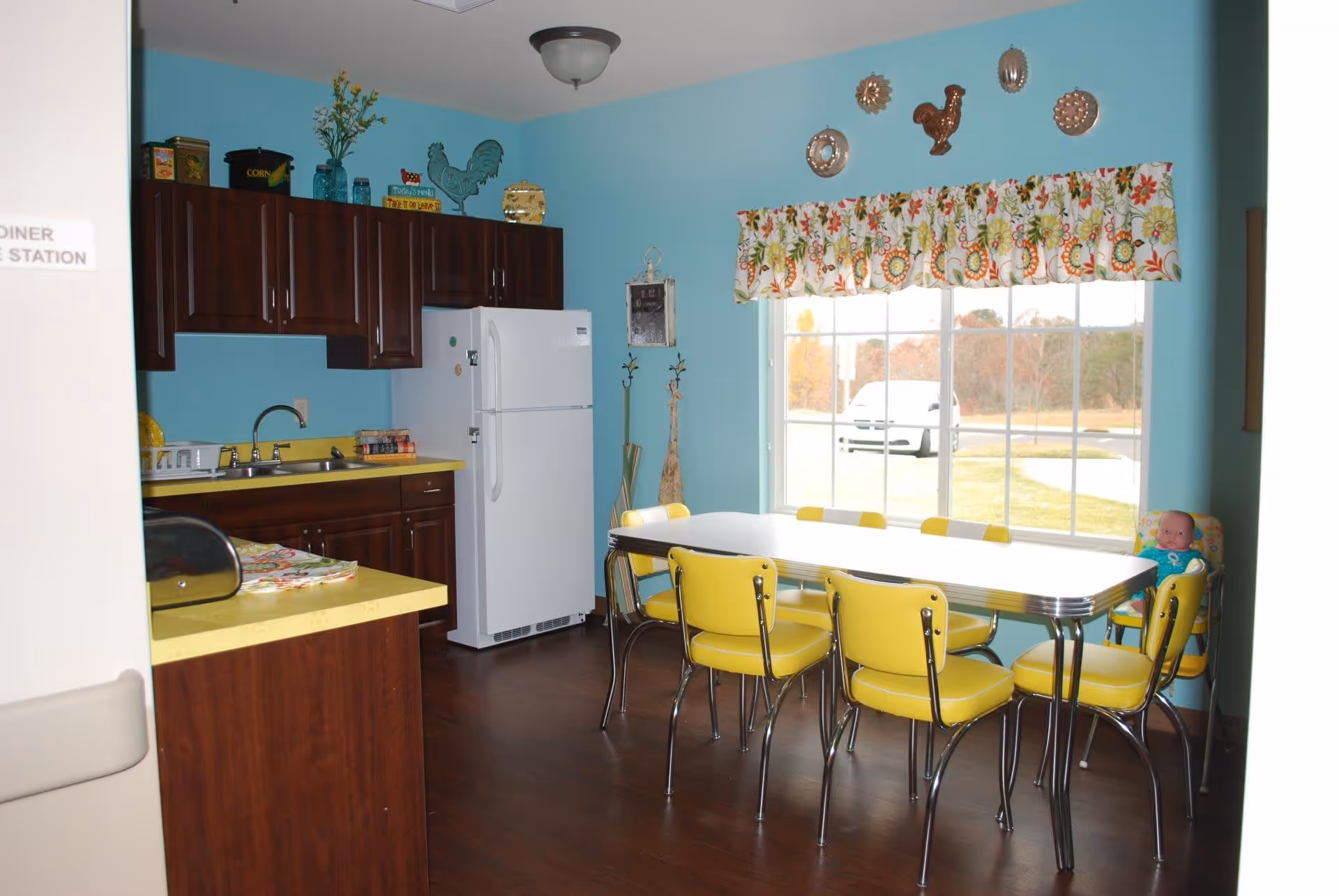 A bright kitchen and dining area with turquoise walls and dark wood cabinets. The kitchen has a white refrigerator, a yellow countertop, and a sink. The dining area features a white table with chrome legs surrounded by yellow chairs. A large window with a floral valance lets in natural light, and decorative plates and a rooster ornament hang on the wall above the window. A doll is seated in a corner chair near the window.