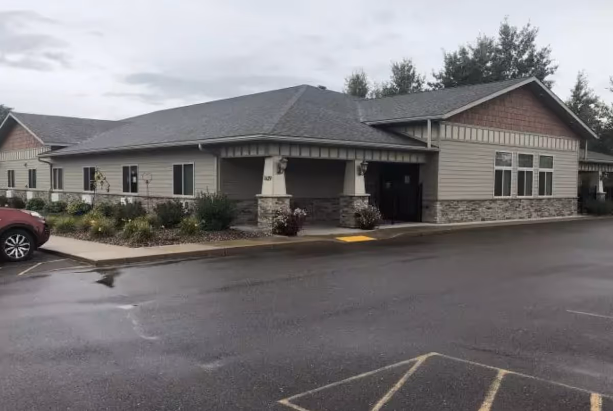 Exterior view of a single-story building with a covered entrance, stone and siding facade, surrounded by some landscaping and a parking lot with a red car parked on the left side.