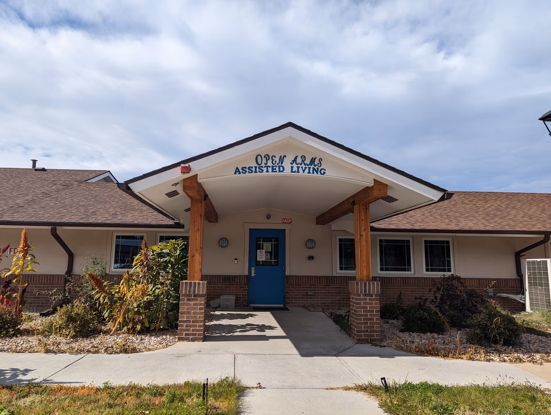 Front exterior view of Open Arms Assisted Living facility showing the entrance with a blue door under a peaked roof supported by wooden beams and brick pillars. There are plants and shrubs on either side of the walkway leading to the door, and the sky is partly cloudy.