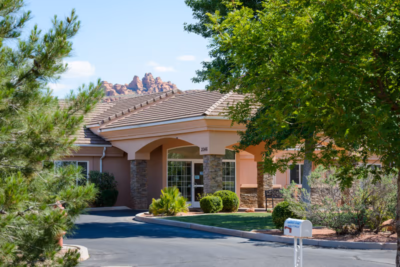 Exterior view of a single-story assisted living facility building with a tiled roof, stone pillars, and a covered entrance. The building is surrounded by green trees and shrubs, with a paved driveway leading to the entrance. Rocky hills are visible in the background under a clear blue sky.