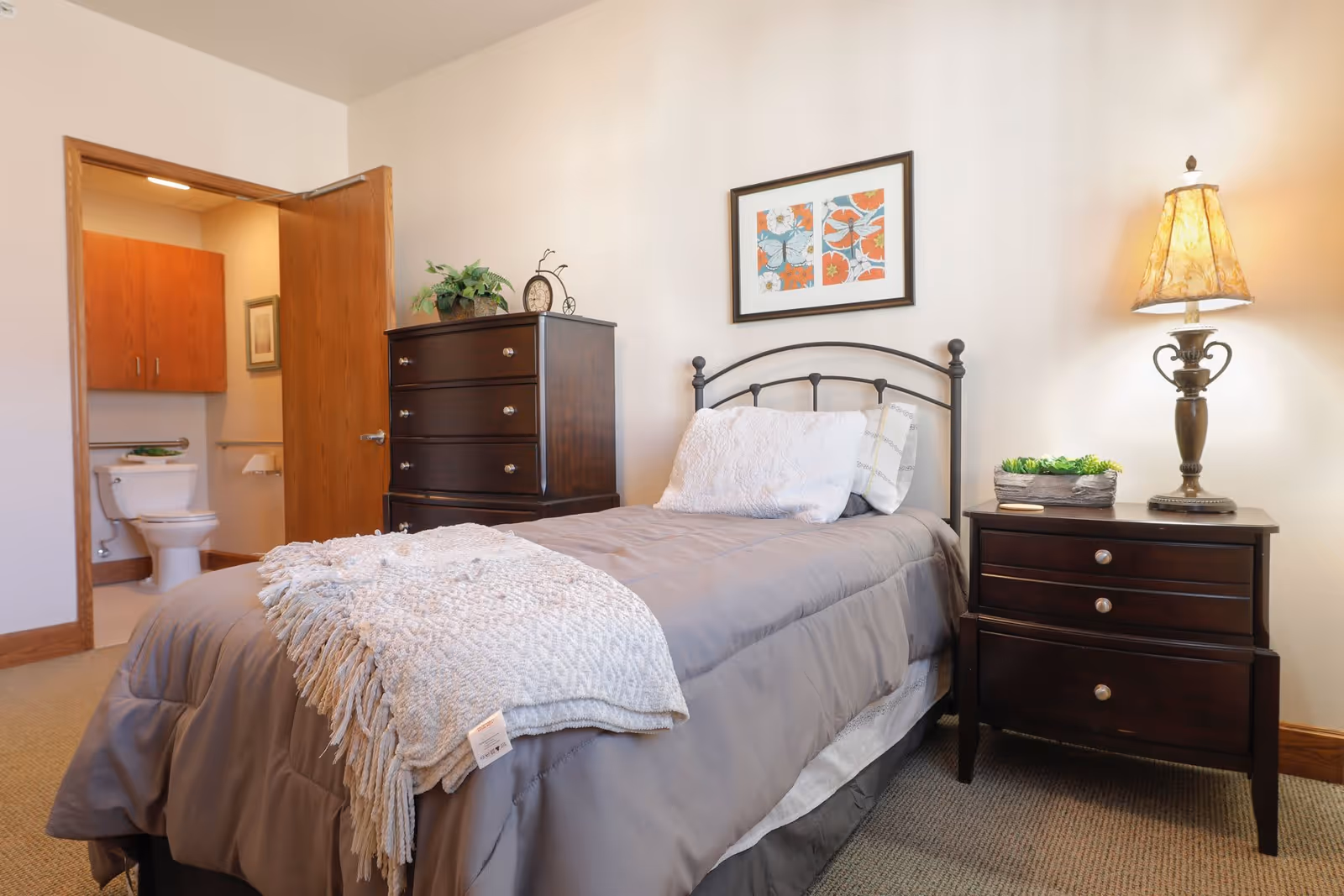A cozy bedroom with a single bed covered in gray bedding and a white textured throw blanket. Next to the bed is a dark wooden nightstand with a decorative lamp and a small plant. On the opposite side, there is a matching dark wooden dresser with a plant and a clock on top. A framed colorful artwork hangs on the wall above the bed. An open door reveals a bathroom with a toilet and wooden cabinets.