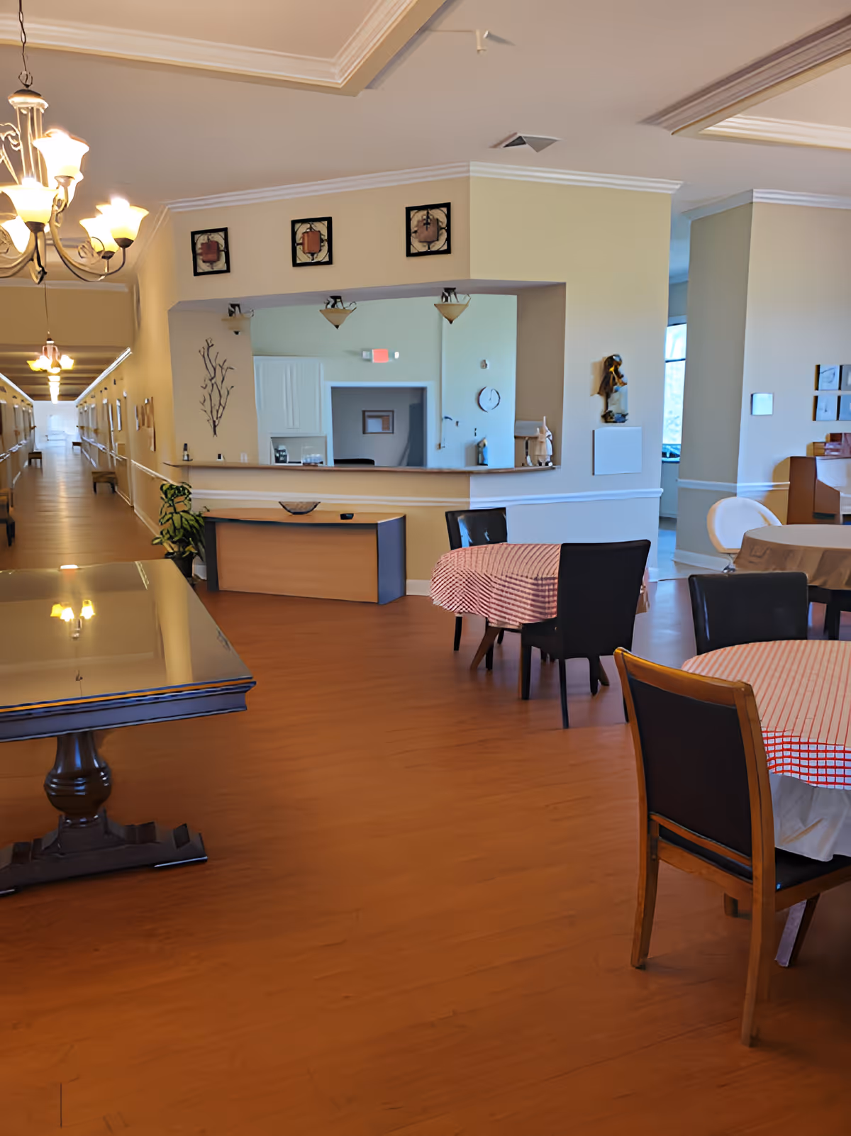 Interior view of a senior care facility dining area with tables covered in red and white checkered tablecloths, black chairs, a wooden floor, and a reception desk in the background. The hallway extends to the left with handrails and lighting fixtures on the ceiling.