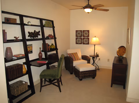 A cozy interior room featuring a dark wooden ladder-style bookshelf with various decorative items and books, a green upholstered chair, a wicker armchair with a matching ottoman and a striped cushion, a side table with a lamp, and a small dark wooden dresser with a decorative plate on top. The walls are light-colored and adorned with framed floral artwork and a ceiling fan with a light fixture.