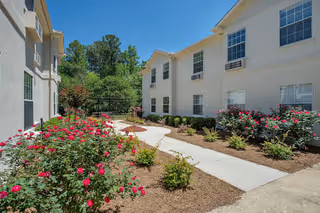Sunlit courtyard walkway between two white residential buildings lined with rose bushes and landscaping.