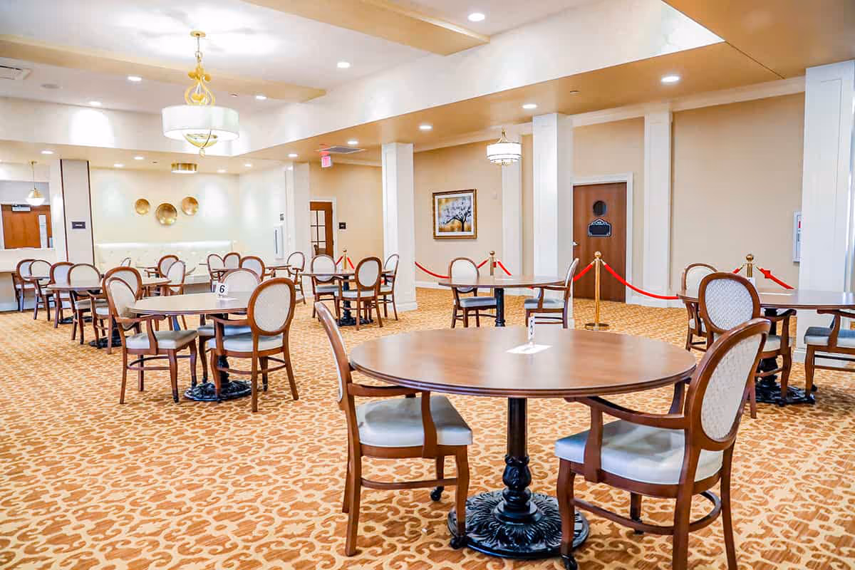 A spacious dining room with multiple round and rectangular wooden tables, each surrounded by cushioned chairs with wooden frames. The room has a patterned carpet, beige walls, and ceiling lights including chandeliers. There are red velvet ropes sectioning off an area near a door, and decorative wall art is visible.