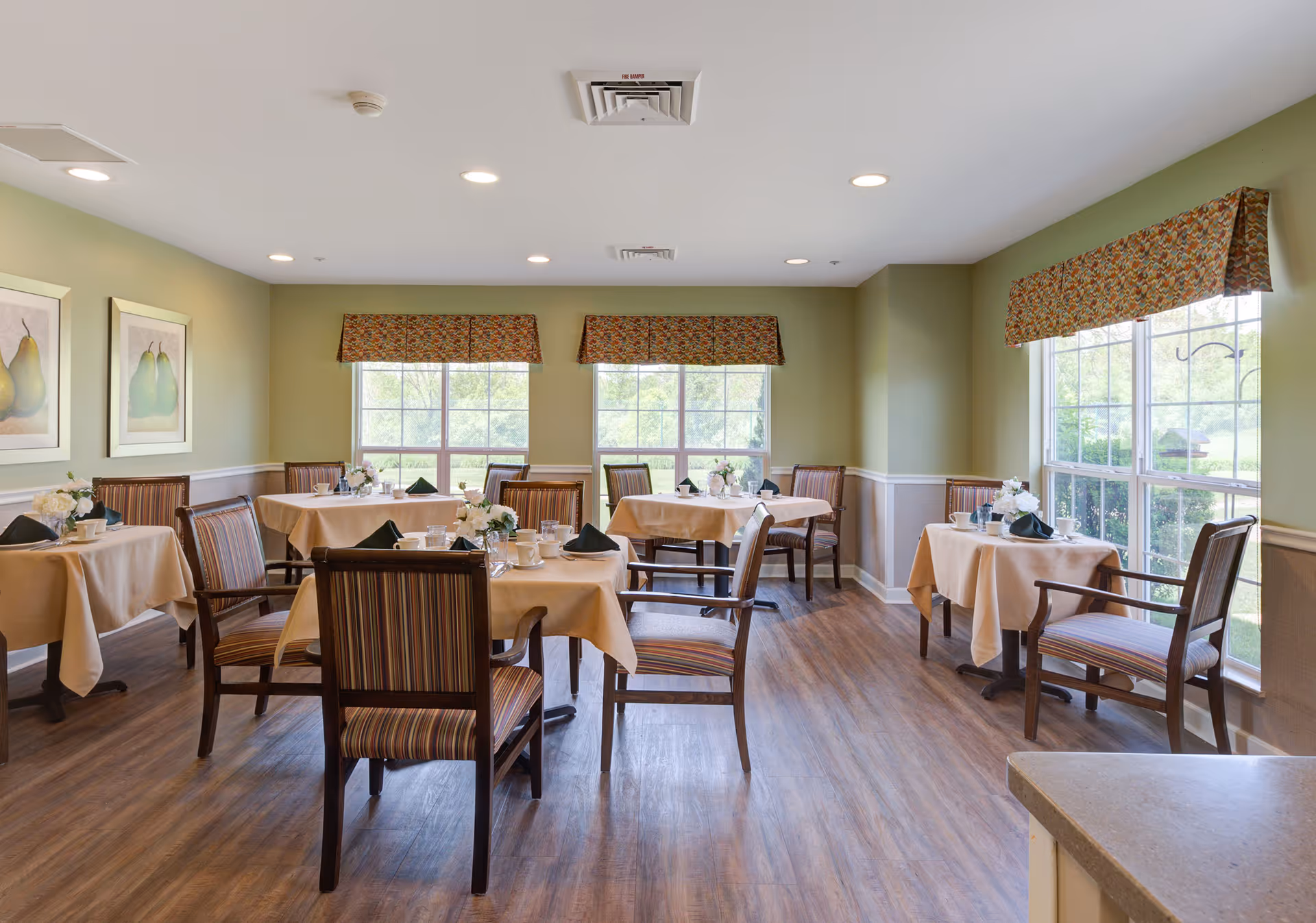 A bright dining room with several tables covered in beige tablecloths, each set with cups, plates, and black napkins. The room has large windows with floral valances letting in natural light, green walls with white trim, wooden chairs with striped cushions, and framed artwork of pears on one wall.
