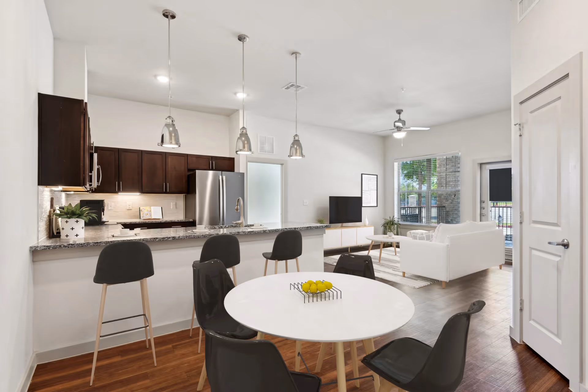 Open-plan interior showing a kitchen with dark cabinets and a breakfast bar, a round dining table, and a white sofa in the living area.