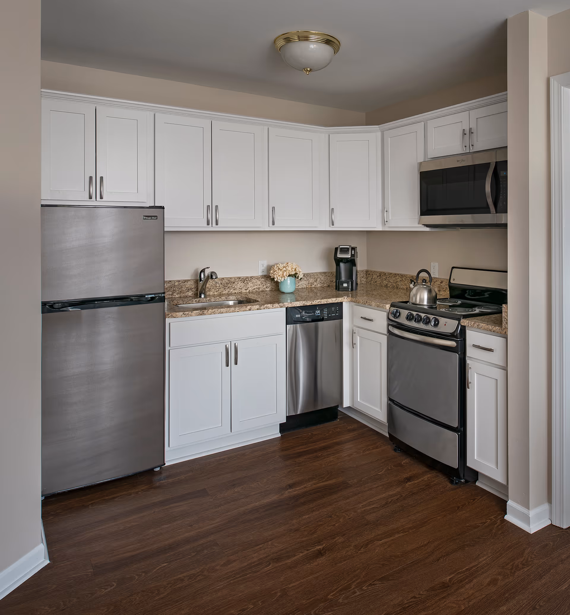 A modern kitchen with white cabinets, stainless steel refrigerator, dishwasher, stove, and microwave. The countertop is granite with a small sink and a coffee maker. The floor is dark wood, and there is a ceiling light fixture.