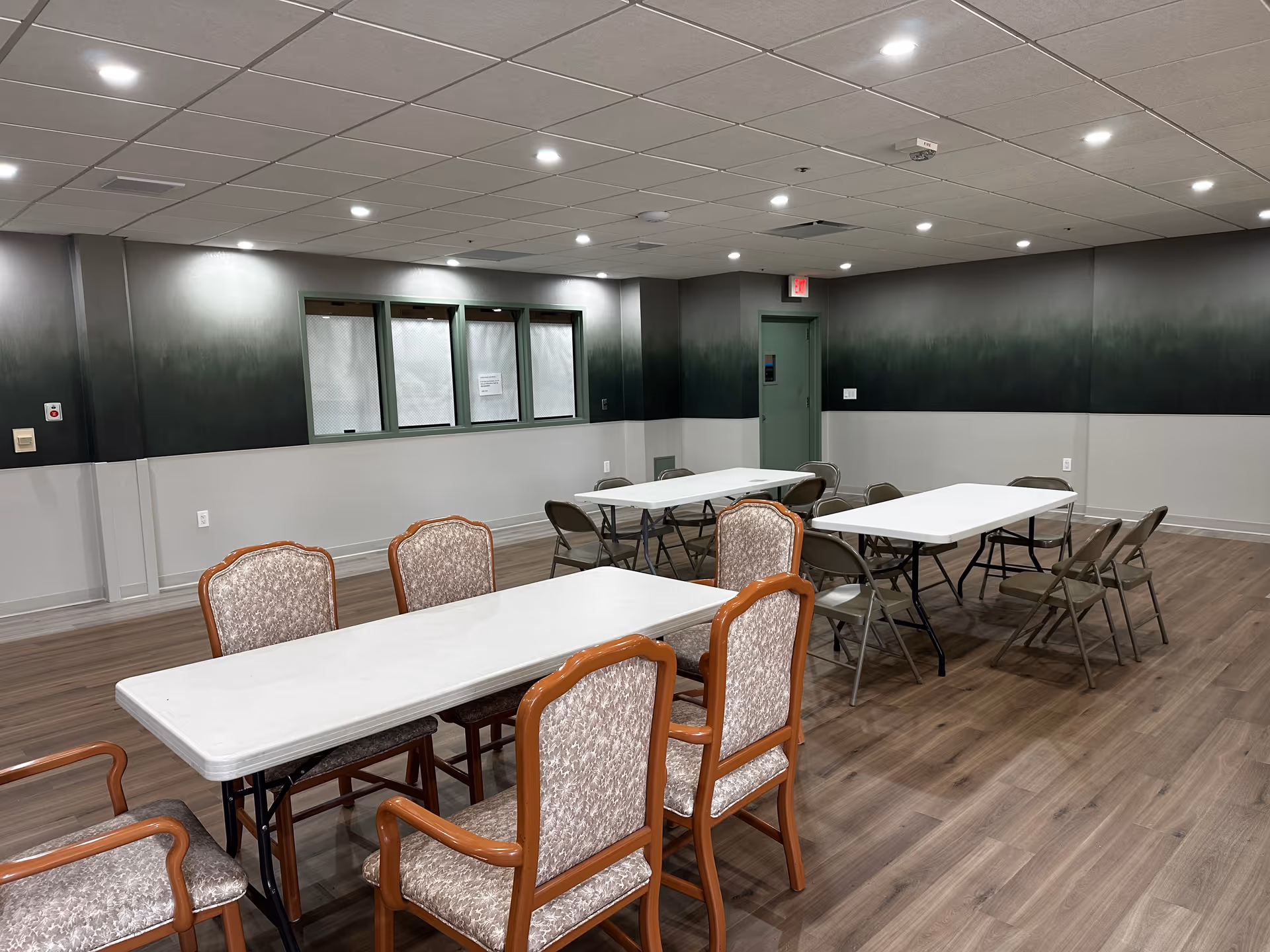 Empty community dining/activity room with folding tables and a mix of upholstered and metal chairs on a wood floor.