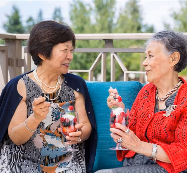 Two elderly women sitting outdoors on a patio or balcony, enjoying desserts with berries in glass cups and engaging in conversation. One woman is wearing a patterned dress with a dark shawl and pearl necklace, while the other is wearing a red knitted cardigan and a necklace.