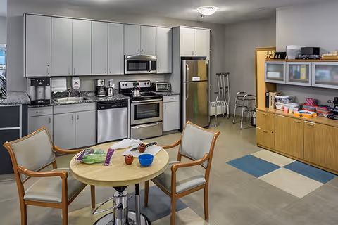 Communal kitchen and dining area with a round table and chairs, stainless steel appliances, cabinets, and mobility aids against the wall.