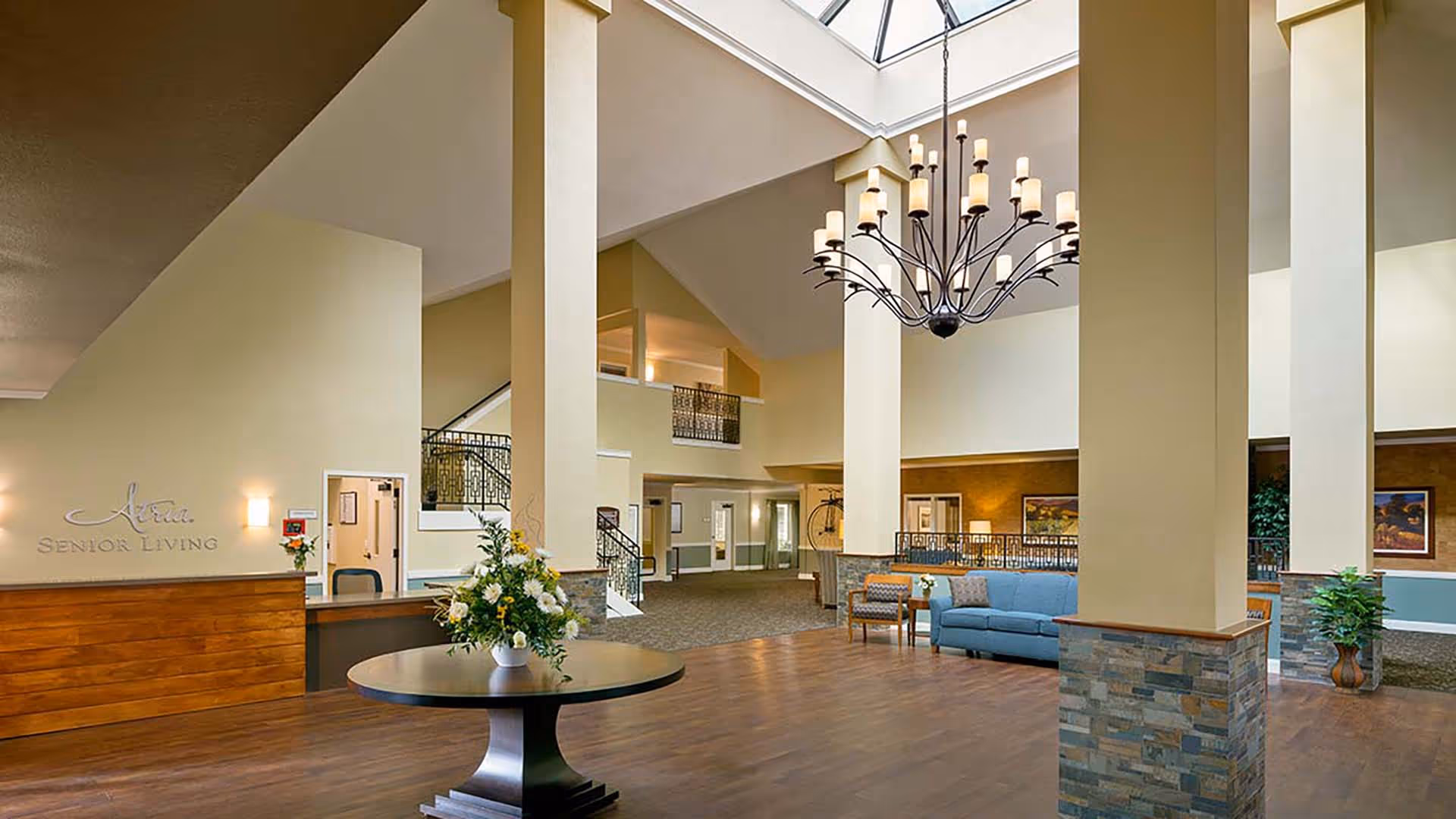 Open senior living lobby with high ceilings, a large chandelier, reception desk, seating area, and a central table with flowers.
