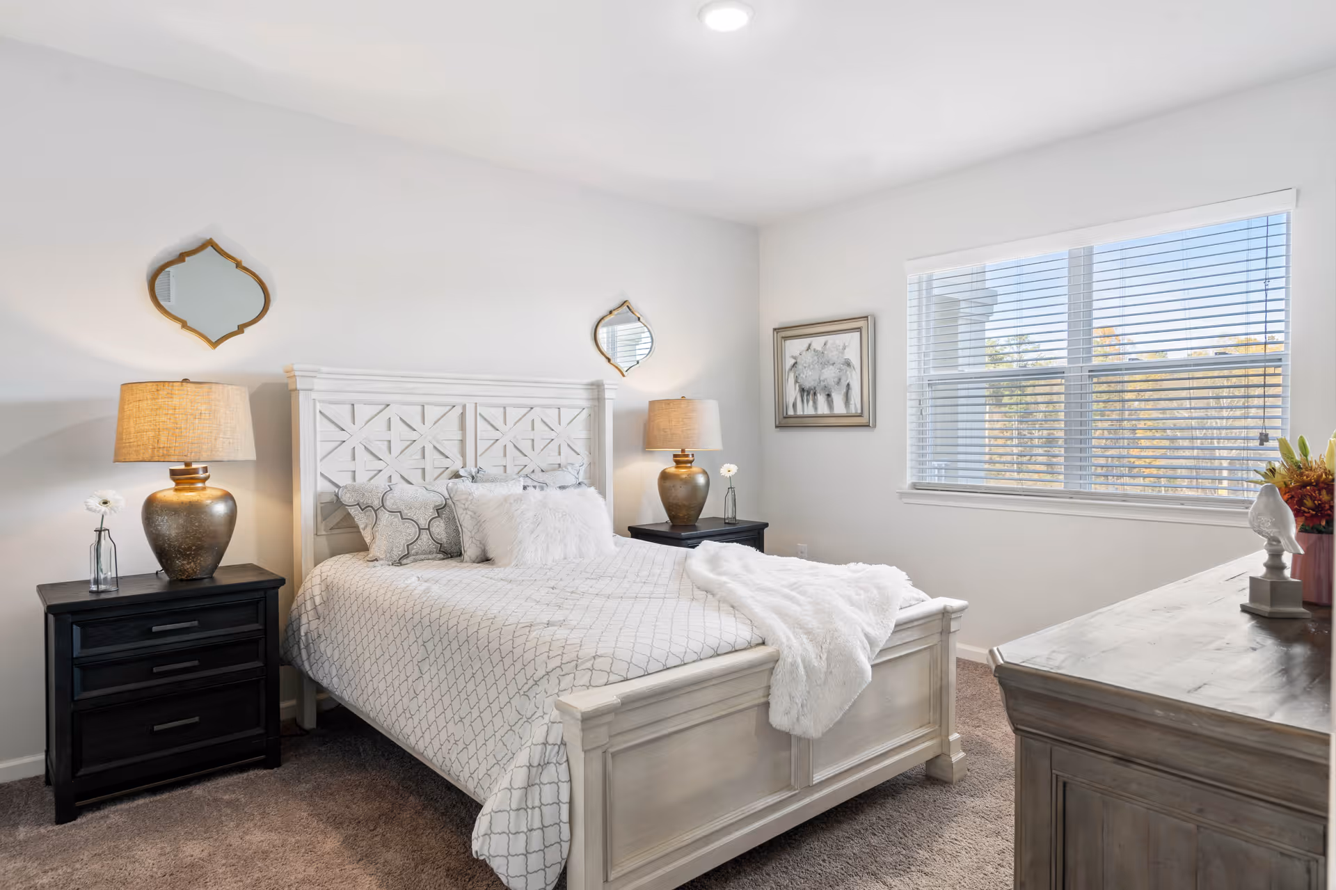 A bright and cozy bedroom featuring a white wooden bed with patterned bedding and multiple pillows. Two matching bedside tables with lamps and small vases with flowers flank the bed. Two decorative mirrors hang above the bedside tables. A large window with white blinds lets in natural light, and a framed artwork is on the wall next to the window. A wooden dresser with decorative items is visible in the foreground.