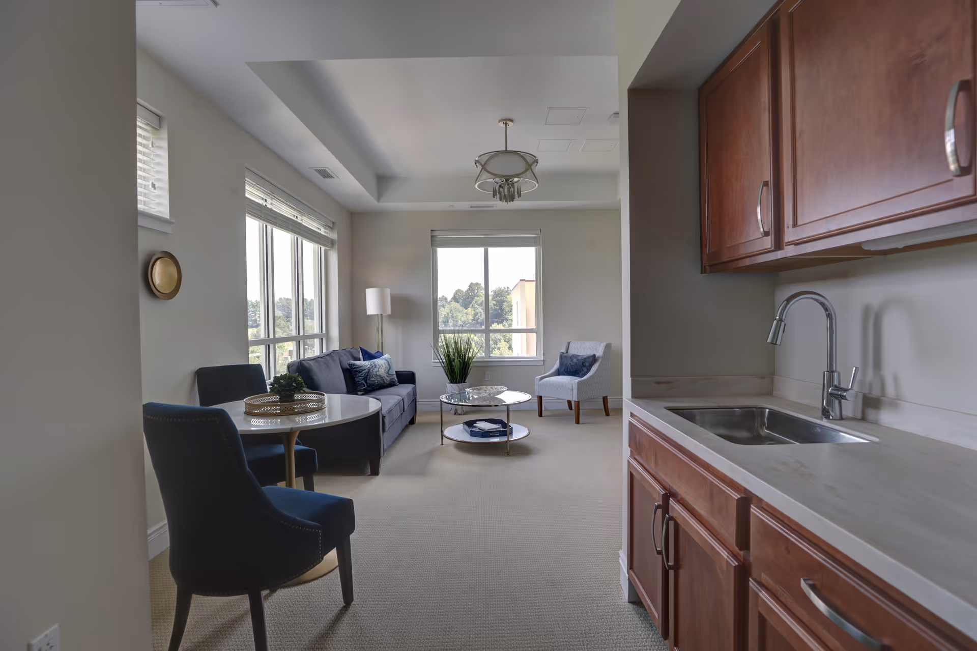 Light-filled living area with a small kitchenette to the right, a sofa and chairs around a coffee table, and large windows.
