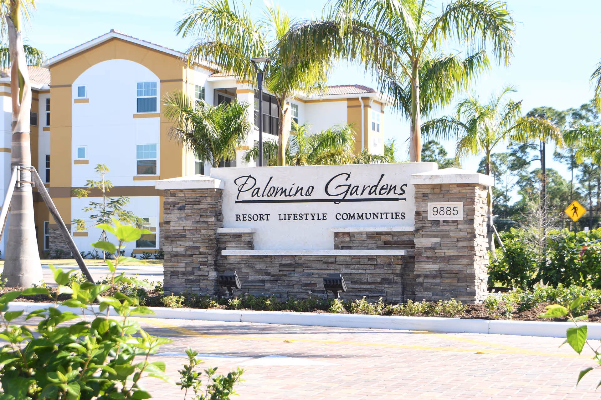 Stone entrance sign reading 'Palomino Gardens Resort Lifestyle Communities' in front of a multi-story building and palm trees.