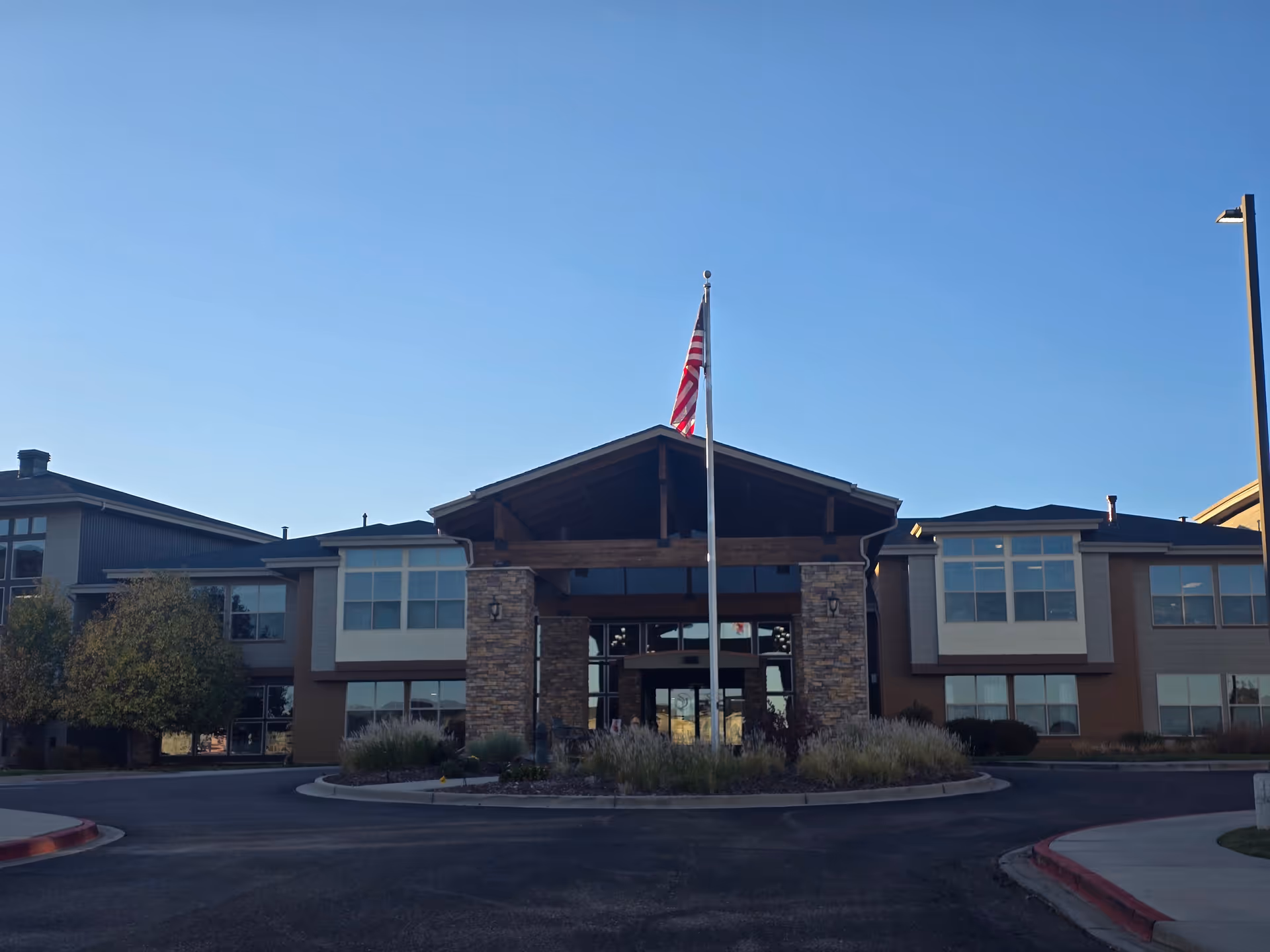 Front exterior view of a two-story senior living facility building with a peaked roof entrance supported by stone pillars. An American flag is flying on a flagpole in front of the entrance. The sky is clear and blue.