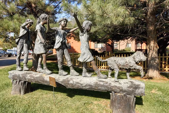 Bronze sculpture of four children and a dog walking in a line on a large log outdoors, with trees and a brick building in the background.