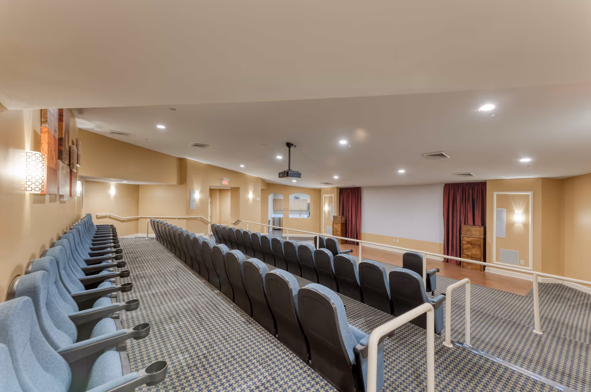 Interior view of a small theater or presentation room with rows of blue cushioned seats arranged in tiers facing a large white projection screen. The room has beige walls, carpeted floors with a patterned design, wall-mounted lights, and a ceiling-mounted projector. There are red curtains on either side of the screen and a small wooden podium to the right.
