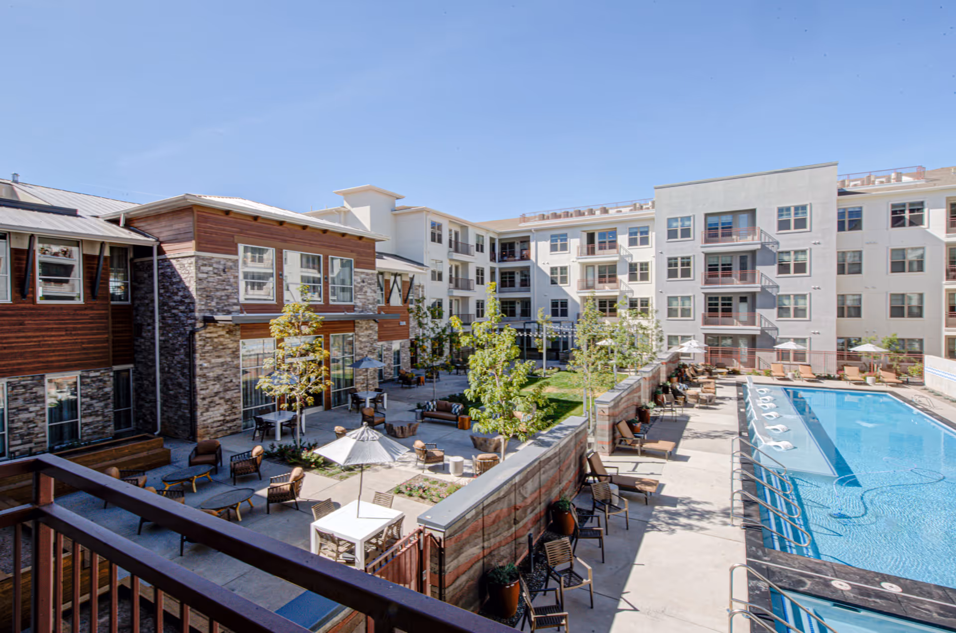 Outdoor courtyard area of a senior living facility featuring a swimming pool with lounge chairs, patio seating with tables and umbrellas, small trees, and multi-story residential buildings surrounding the courtyard under a clear blue sky.