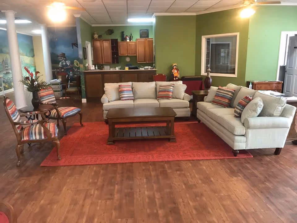 Communal living room with two sofas, striped chairs and a coffee table on a red rug with a small kitchenette/bar behind.