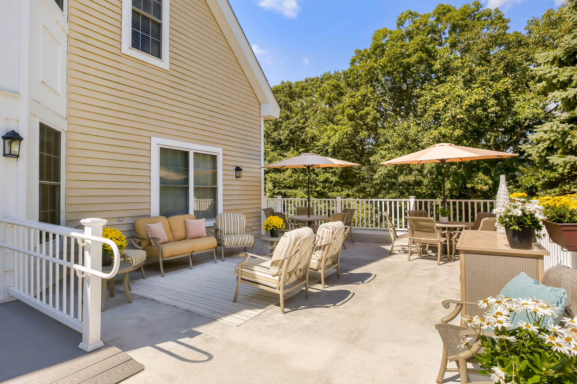 Outdoor patio area with beige siding building on the left, featuring cushioned seating, striped chairs, tables with umbrellas, potted plants, and surrounded by green trees under a blue sky.