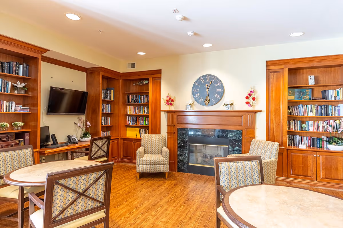 A cozy common area with wooden bookshelves filled with books, a wall-mounted TV, a computer desk with a phone, a fireplace with a large clock above it, and several patterned armchairs and round tables with chairs on a wooden floor.
