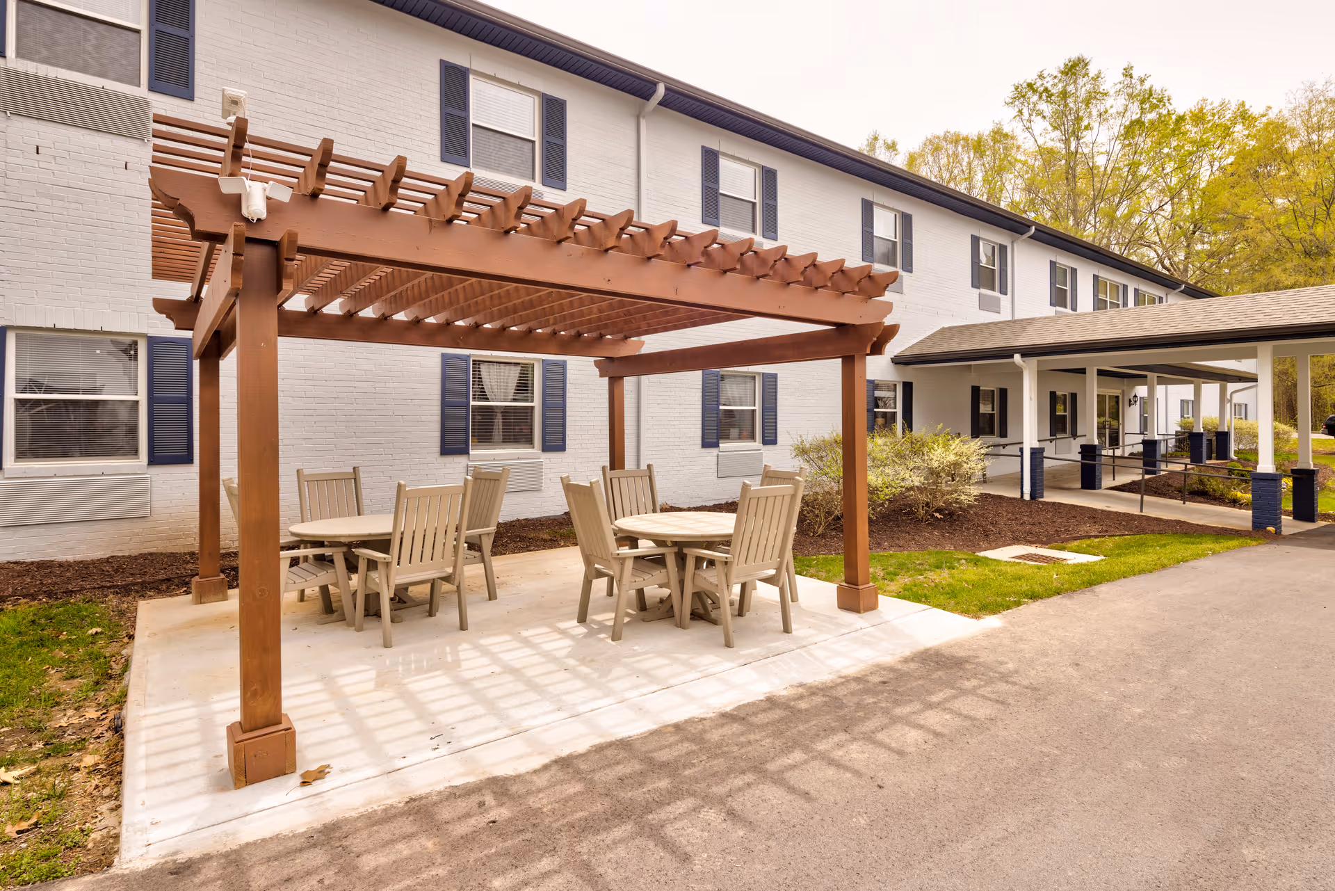 Outdoor seating area with two round tables and several chairs under a wooden pergola next to a two-story white brick building with blue shutters and a covered entrance.