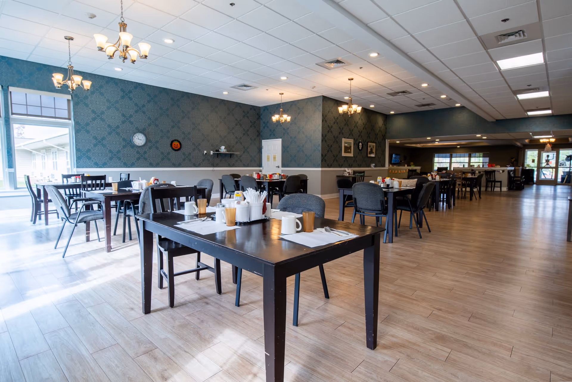 A spacious dining room with several dark wooden tables and gray cushioned chairs. Each table is set with placemats, cups, utensils, and condiment holders. The room has light wood flooring, teal patterned wallpaper, and multiple ceiling lights including chandeliers. Large windows on the left side allow natural light to fill the space.