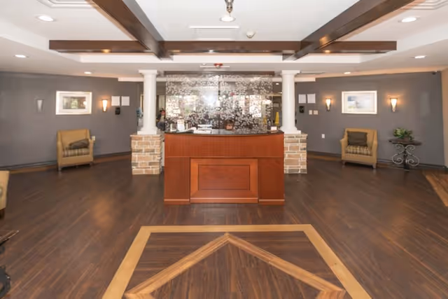 Interior view of a reception area in a senior living facility with a wooden reception desk centered between two white columns with brick bases. The room has dark wood flooring with a decorative inlay, gray walls, framed artwork, wall sconces, and two armchairs with cushions placed against the walls.