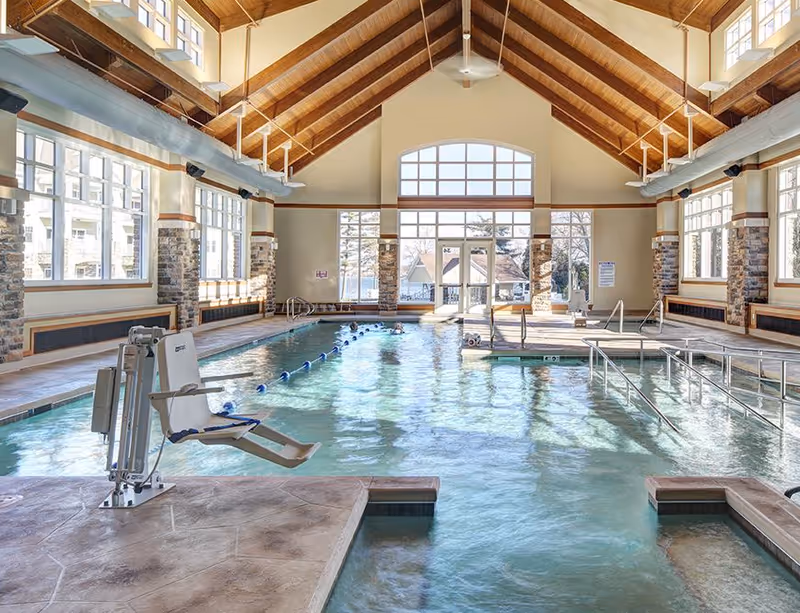 Indoor swimming pool with a lift chair, lap lane ropes, handrails and a high wood‑beamed vaulted ceiling with large windows.