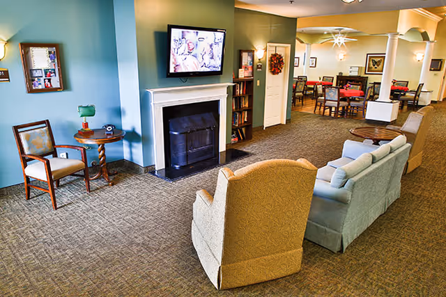 Communal lounge with armchairs and a sofa facing a wall-mounted TV above a fireplace, with dining tables visible in the background.