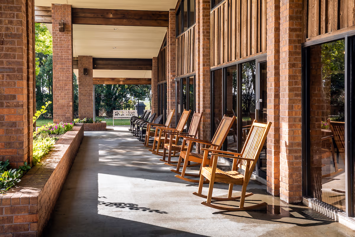 A covered outdoor patio area with a row of wooden rocking chairs lined up against large glass windows. The patio has brick pillars and a brick planter with green plants and flowers. Sunlight casts shadows on the concrete floor.