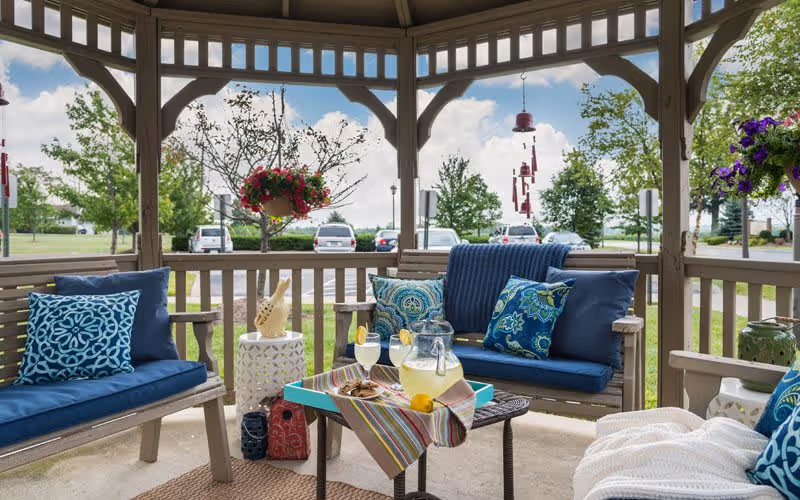 Gazebo-style outdoor seating area with blue-cushioned benches, patterned pillows, a small table holding a pitcher of lemonade and glasses, and hanging flower baskets overlooking a parking area.