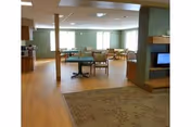 Spacious common area in an assisted living facility with wooden flooring, several chairs and tables arranged for seating, large windows allowing natural light, and a fireplace with a television mounted above it.