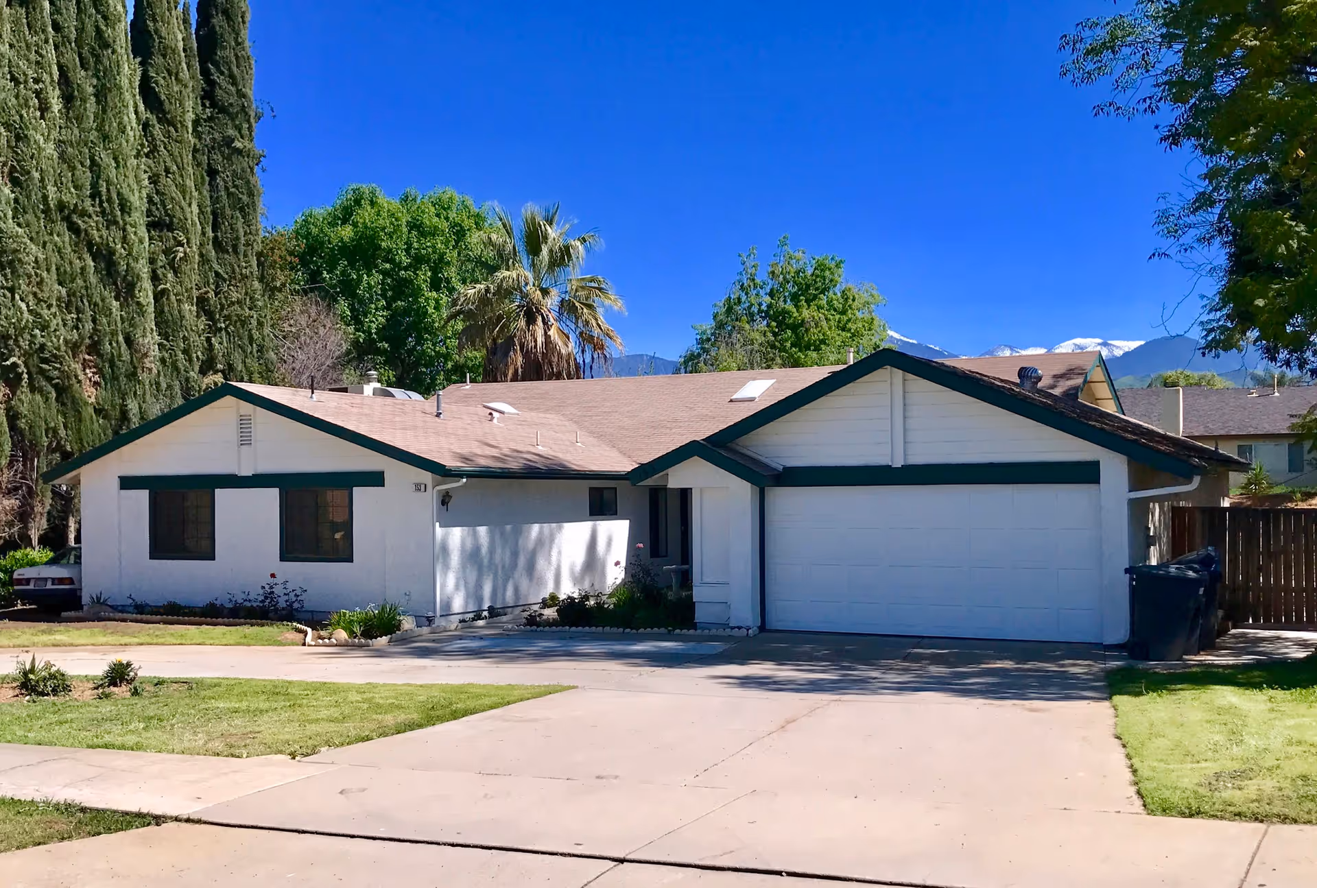 Single-story white ranch-style house with green trim and attached two-car garage, driveway and trees under a clear blue sky.