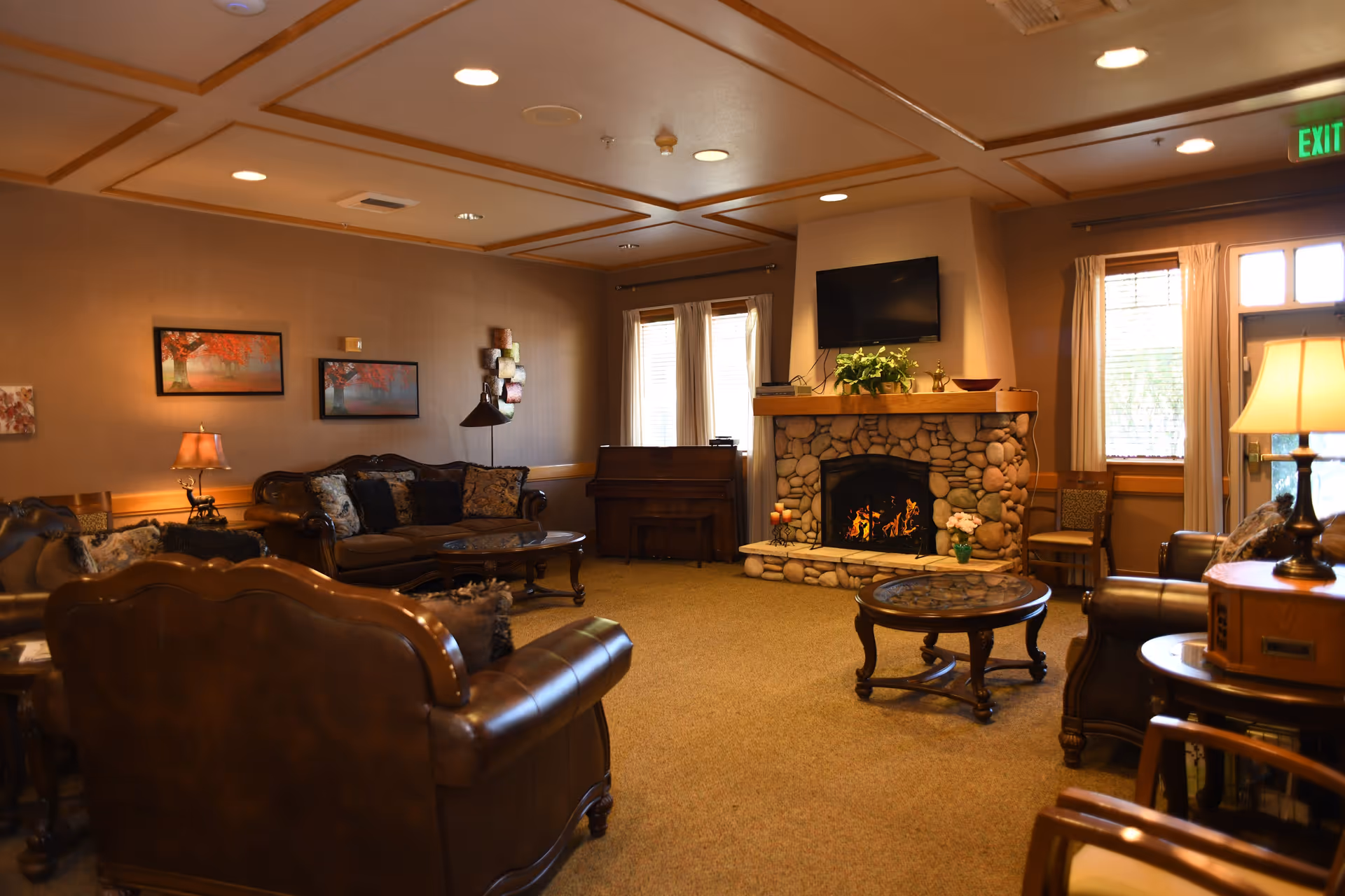 A cozy living room in Oakdale Heights Assisted Living featuring brown leather sofas and armchairs arranged around a stone fireplace with a fire burning. A flat-screen TV is mounted above the fireplace. The room has warm lighting, framed artwork on the walls, a piano near the windows with curtains, and several wooden tables with lamps and decorative items.