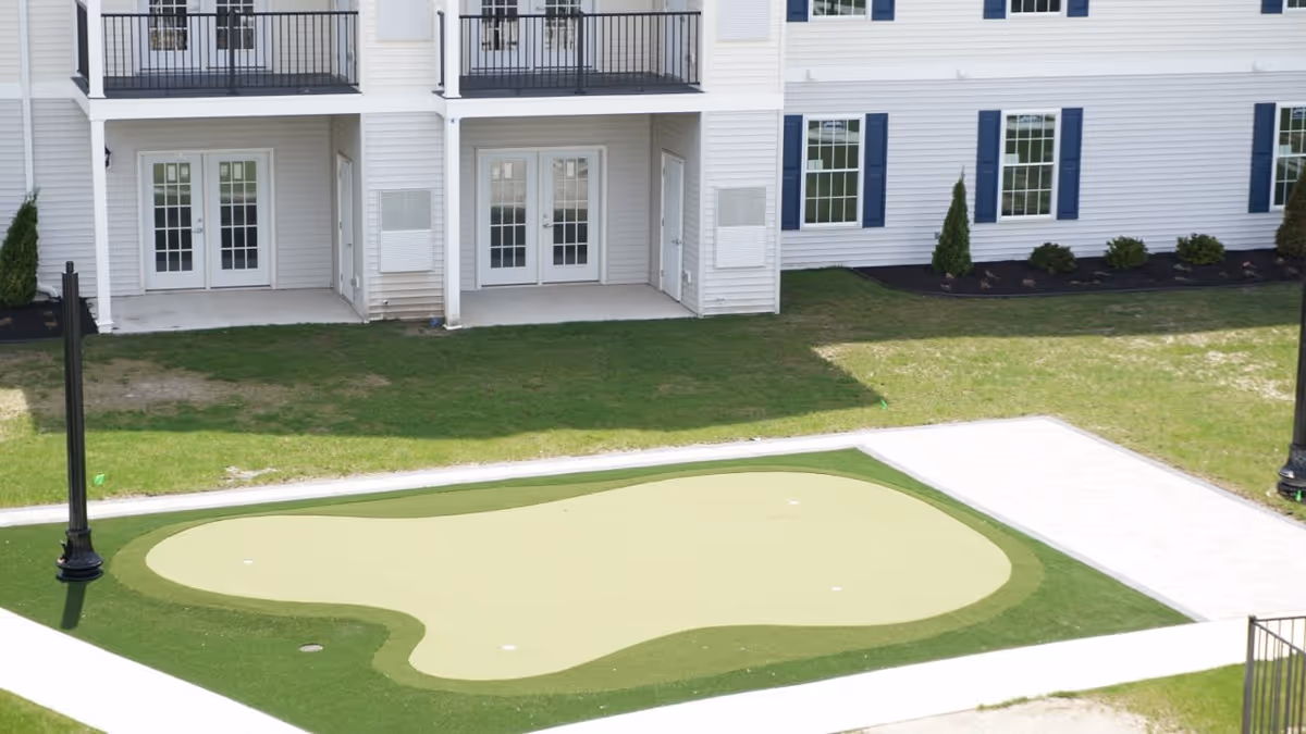 Outdoor putting green area with artificial turf surrounded by a paved walkway, located in front of a white residential building with balconies and multiple glass doors and windows.