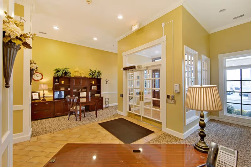 Interior view of a senior living facility lobby with a wooden desk and lamp in the foreground, a glass double door entrance, a wooden cabinet with plants and decorative items, and a window showing the outside parking area.