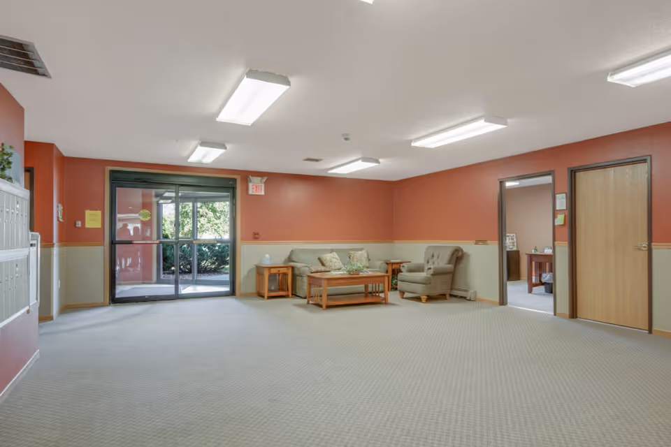 A spacious lobby area with beige carpet and walls painted in two tones of red and beige. There is a seating area with a sofa, armchair, coffee table, and side tables with decorative items. Glass double doors lead outside, and there are two wooden doors and an open doorway on the right side of the room. Fluorescent ceiling lights illuminate the space.