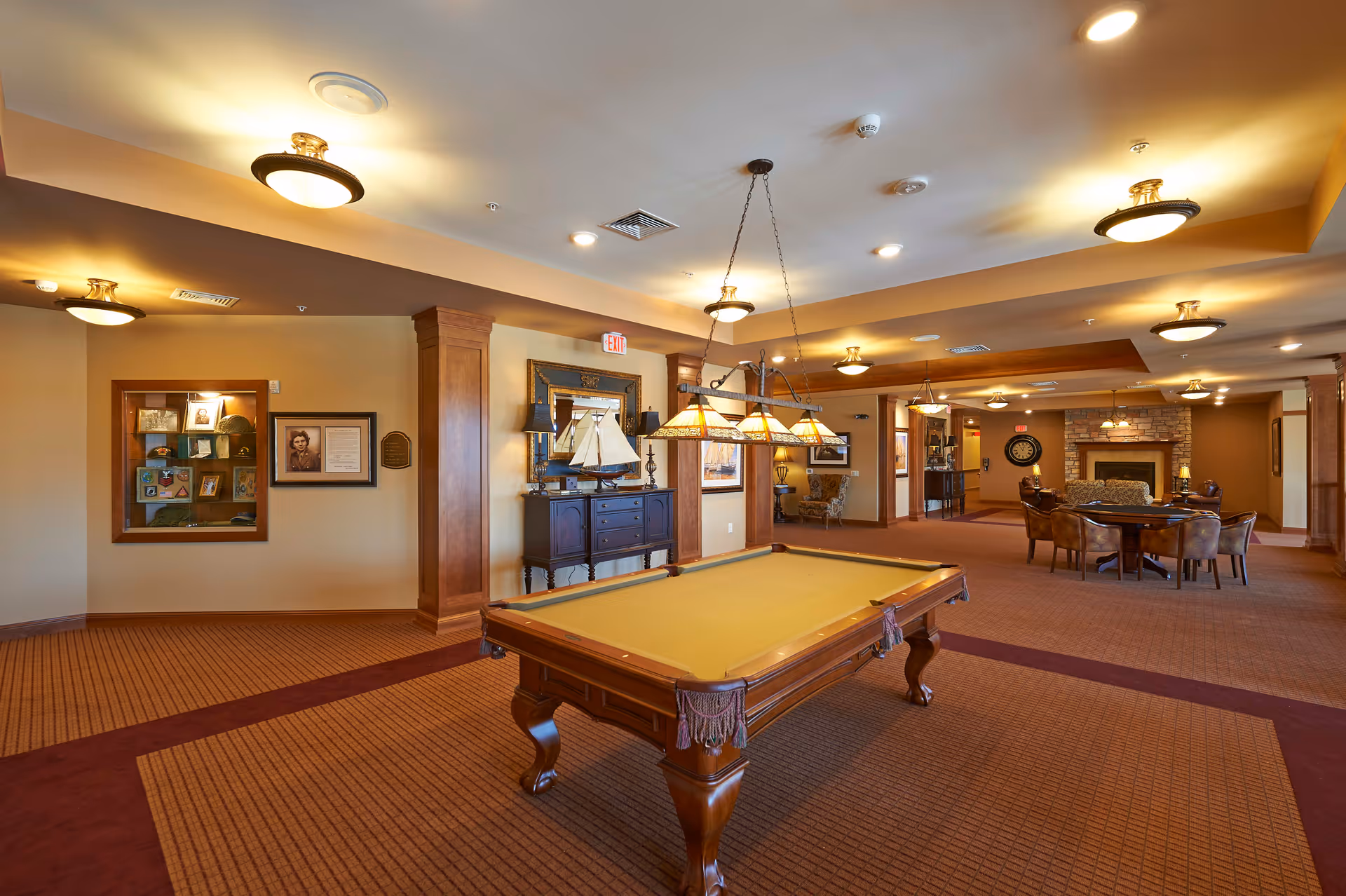 A spacious senior living common area featuring a pool table in the foreground with overhead lighting, a seating area with a round table and chairs, and a fireplace in the background. The room has warm lighting, carpeted floors, and wood accents on the walls and columns.