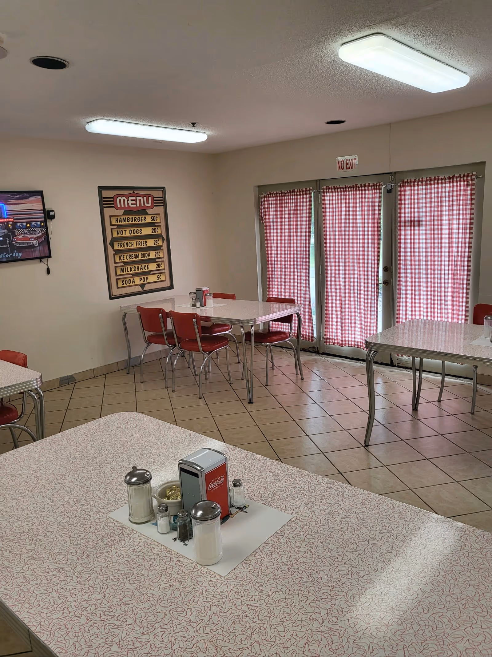 Interior view of a dining area with retro-style tables and red chairs. The tables have vintage patterned surfaces and are set with condiments including salt, pepper, sugar, and a Coca-Cola napkin dispenser. The room has tiled floors, fluorescent ceiling lights, and red and white checkered curtains covering glass doors. A vintage menu sign on the wall lists prices for hamburger, hot dogs, french fries, ice cream soda, milkshake, and soda pop.