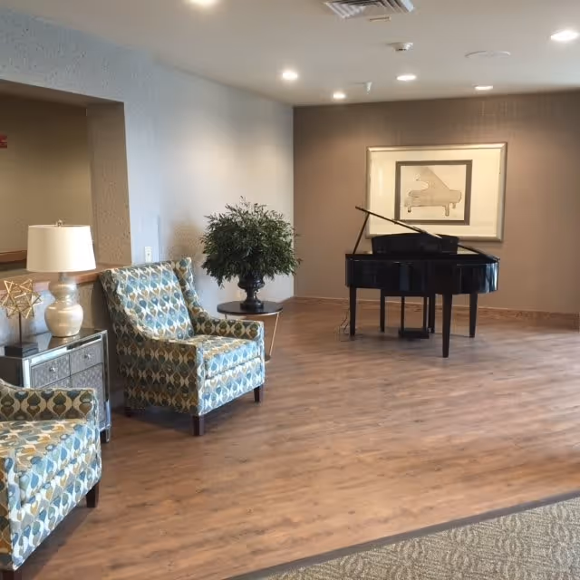 A lounge area with a black grand piano, patterned armchairs, side table with a lamp and a potted plant on wood flooring.