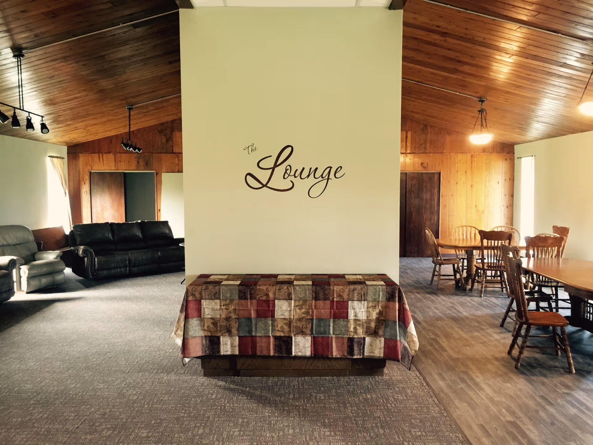 Interior communal lounge with sofas on the left, dining tables on the right, and a central wall labeled "The Lounge" under a wooden ceiling.