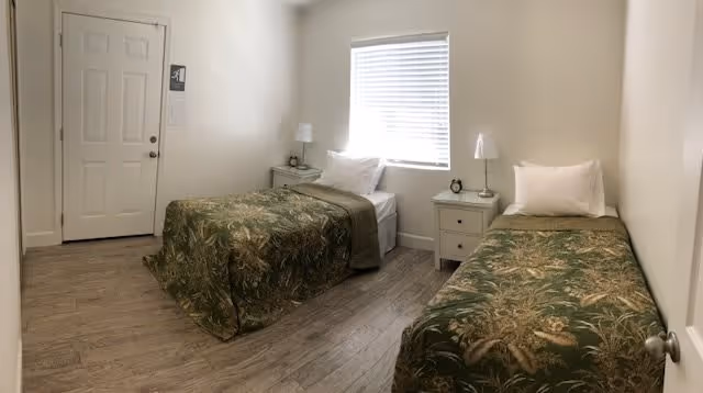 A simple bedroom with two single beds each covered with green patterned bedspreads. Between the beds is a small white nightstand with a lamp and an alarm clock. There is a window with closed blinds on the wall behind the beds. The room has light-colored walls and wood flooring. A closed door is visible on the left side of the room.