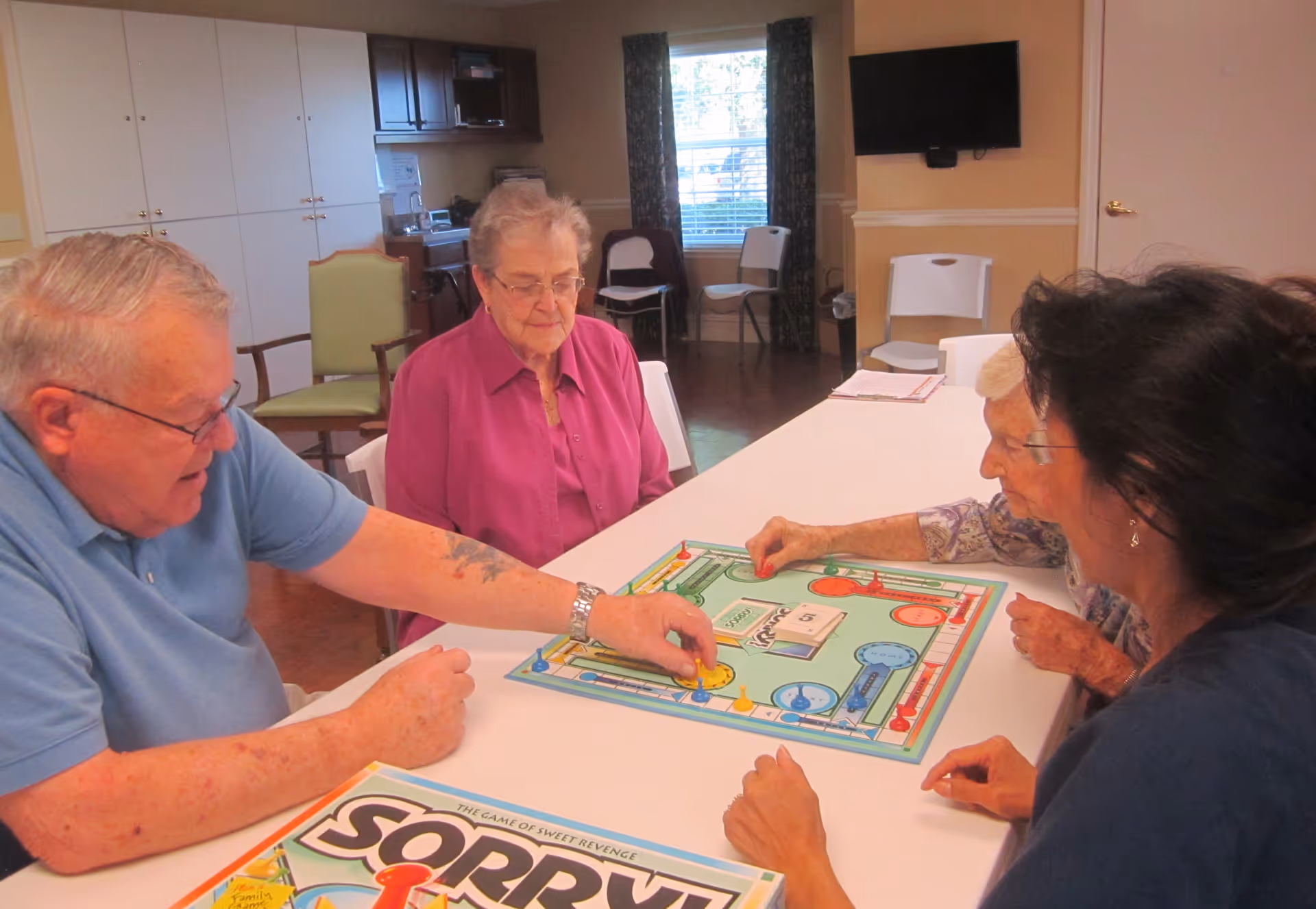 Four elderly people sitting around a table playing the board game Sorry! in a well-lit room with cabinets, chairs, and a wall-mounted TV.