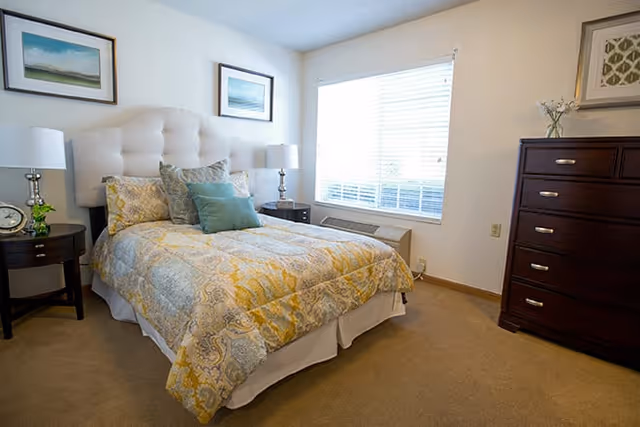 A cozy bedroom with a bed featuring a tufted headboard, patterned yellow and gray bedding, and multiple pillows. There are two bedside tables with lamps, a window with blinds letting in natural light, a dark wooden chest of drawers, and framed artwork on the walls.