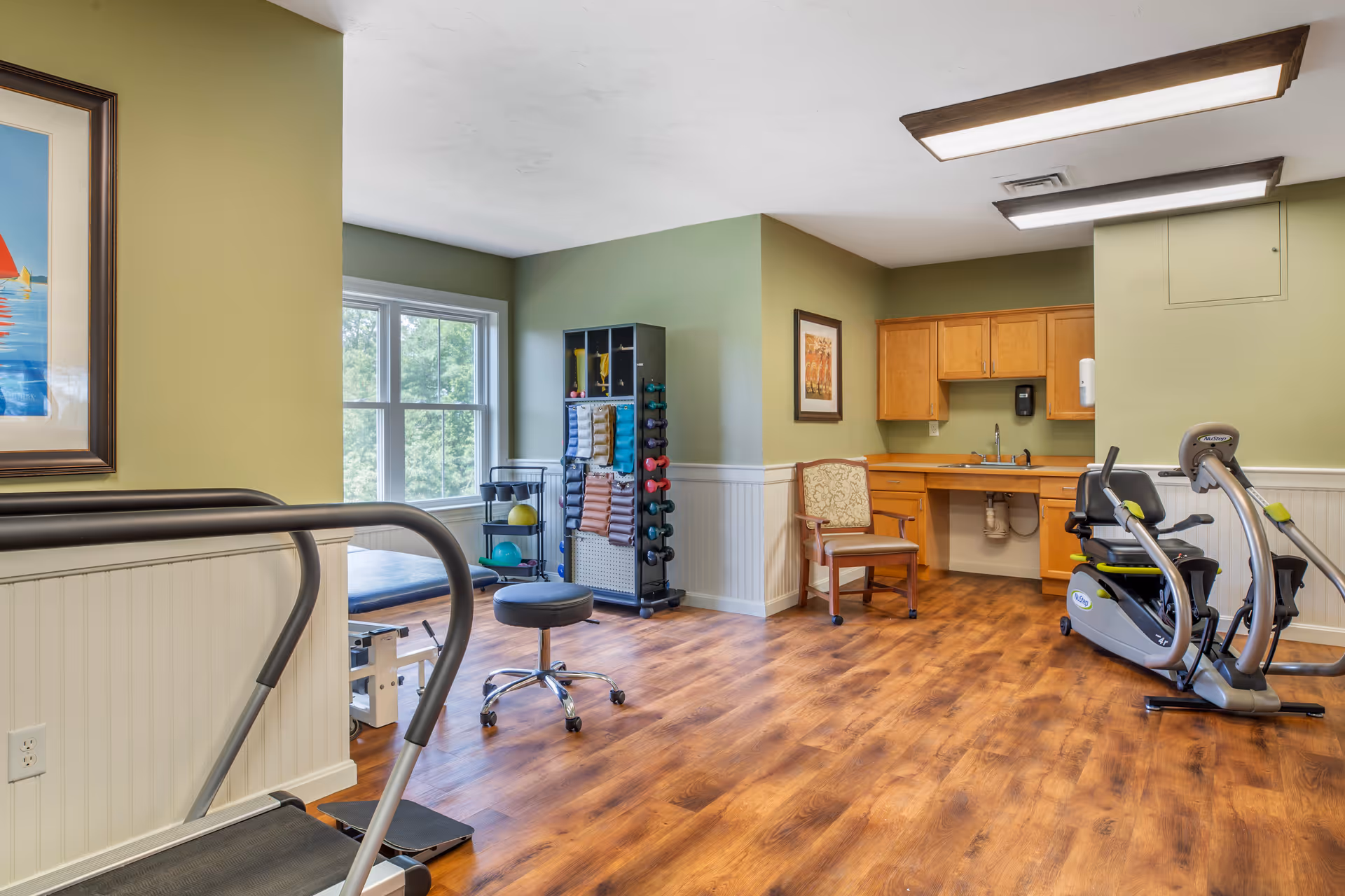 A well-lit exercise room with wooden flooring and light green walls. The room contains various fitness equipment including a treadmill, a recumbent exercise bike, a rack with colorful dumbbells and exercise mats, a padded exercise table, and a rolling stool. There is a wooden chair near a countertop with cabinets and a sink. A large window lets in natural light, and framed artwork is hung on the walls.