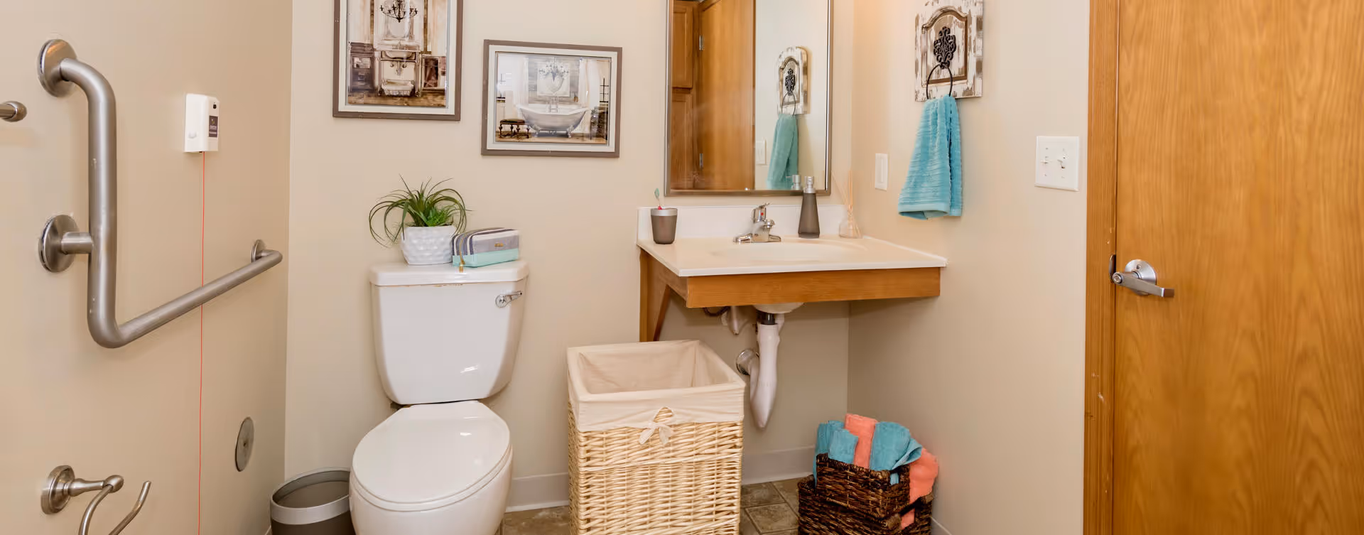 A clean and accessible bathroom featuring a white toilet with a plant and a small radio on top, a wall-mounted sink with a mirror above it, a wicker laundry basket, and a basket with folded towels. The walls are decorated with framed pictures and a turquoise hand towel hangs on a hook. There are grab bars installed near the toilet for accessibility.