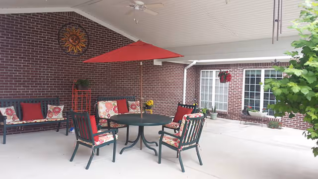 Covered outdoor patio area with a round table and four chairs with floral cushions, a red umbrella, a bench with red and floral cushions, potted plants, a hanging flower basket, and a decorative sun wall ornament on a brick wall.